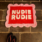 Poppy Nudie Rudie bath mat with pink scalloped border and red centre on stone tile floor, overhead shot