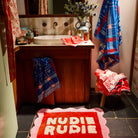 Poppy Nudie Rudie bath mat in red and pink on dark tile floor in a styled bathroom setting