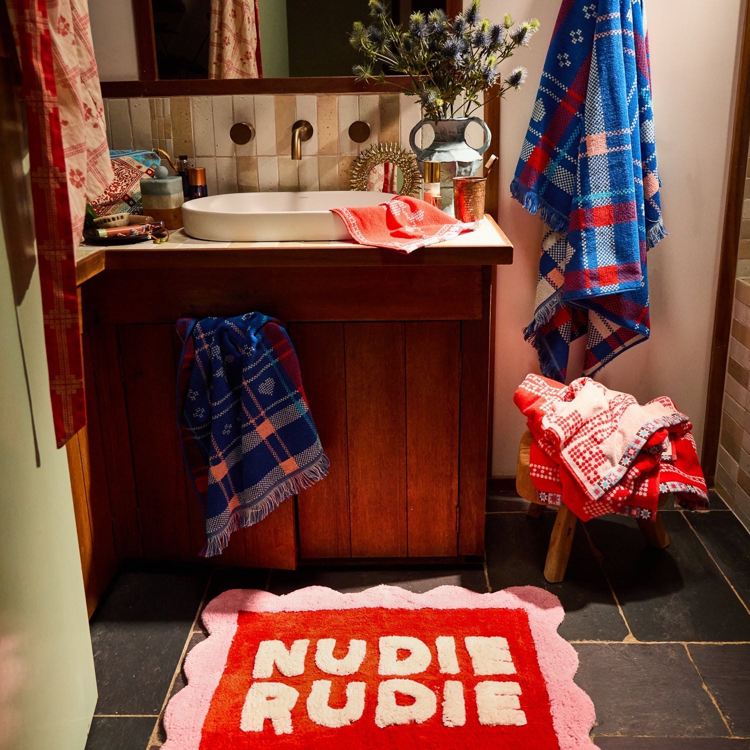 Poppy Nudie Rudie bath mat in red and pink on dark tile floor in a styled bathroom setting