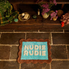Turquoise and brown scallop-edged Nudie Rudie bath mat laid on stone floor beside a wooden bench with bathroom accessories