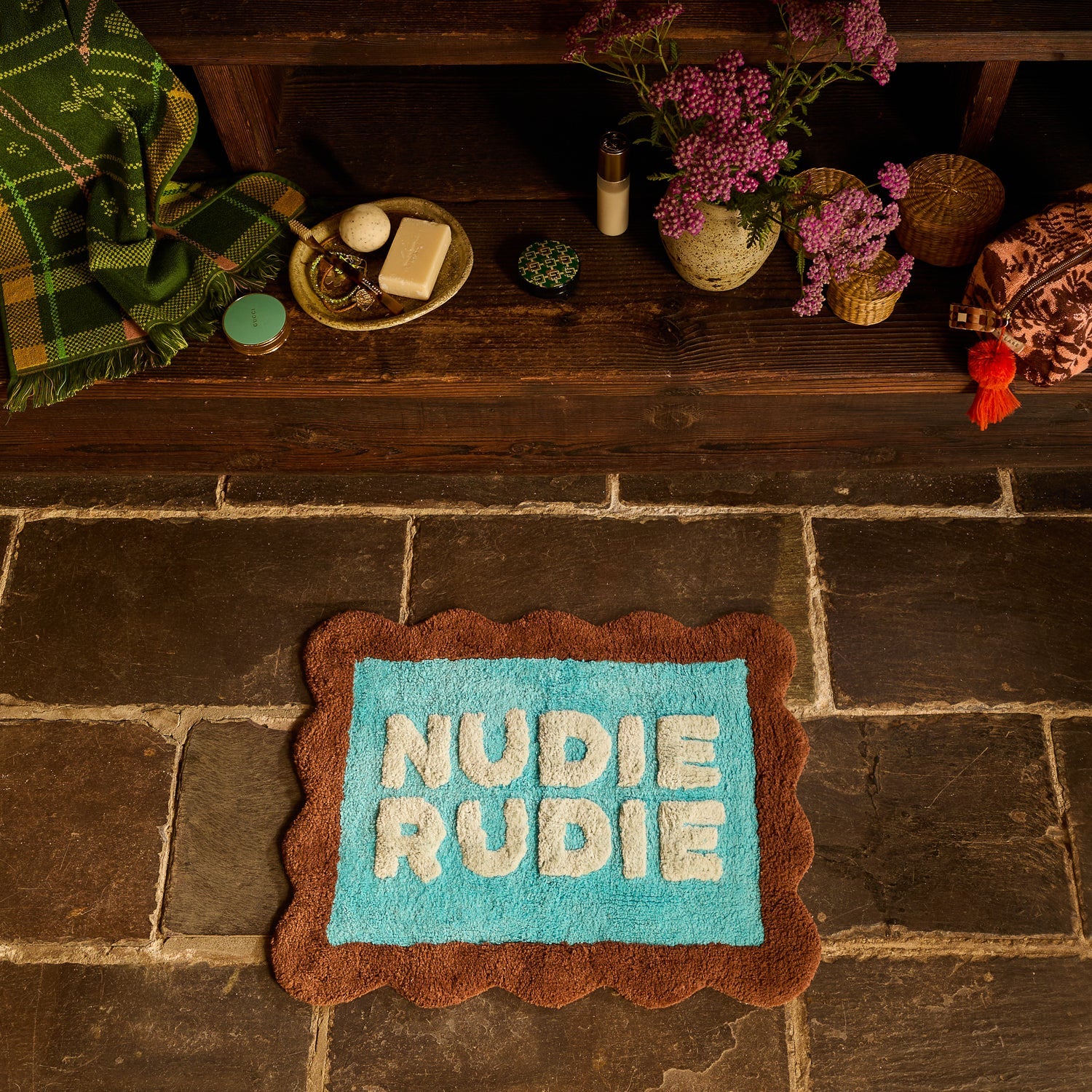 Turquoise and brown scallop-edged Nudie Rudie bath mat laid on stone floor beside a wooden bench with bathroom accessories