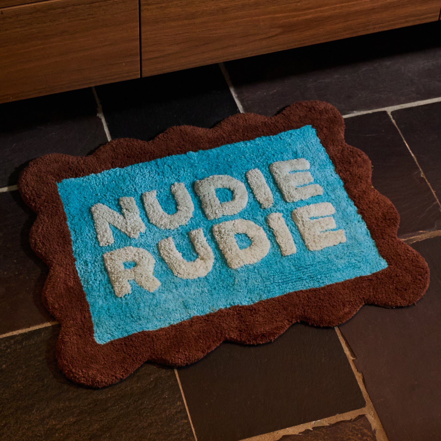 Turquoise tufted Nudie Rudie bath mat with brown scalloped edge on dark slate bathroom floor