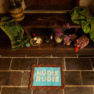Overhead view of turquoise Nudie Rudie bath mat with brown scalloped border on stone floor next to wooden steps