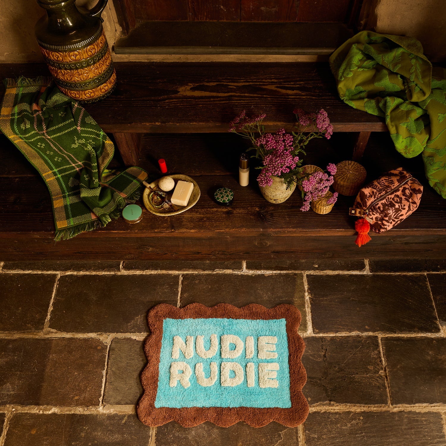 Overhead view of turquoise Nudie Rudie bath mat with brown scalloped border on stone floor next to wooden steps