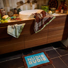 Turquoise Nudie Rudie bath mat with brown scalloped border placed in front of a wooden bathroom vanity