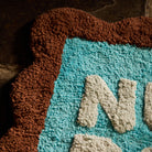 Close-up detail of the tufted texture and scalloped edge on a turquoise and brown bath mat with cream lettering