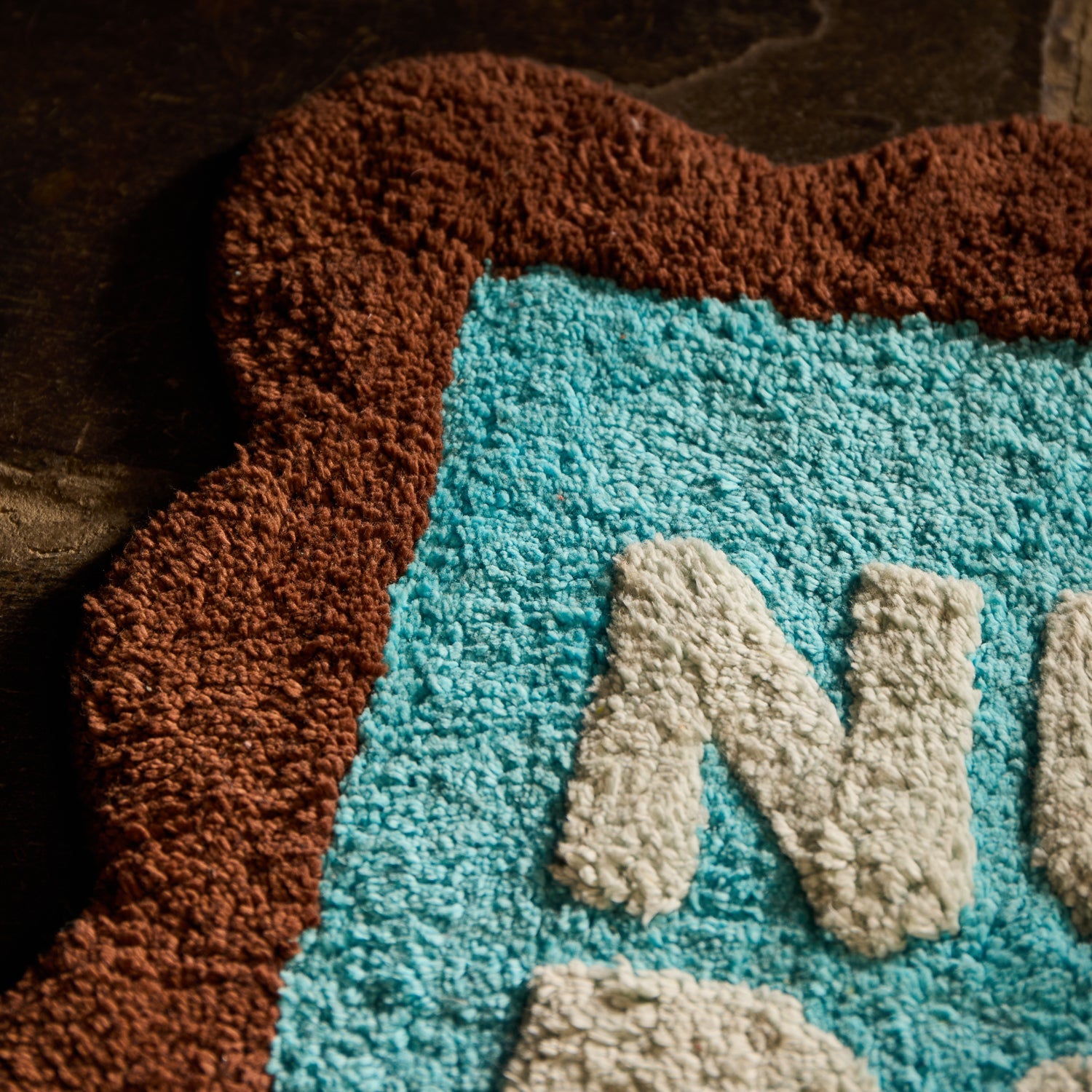 Close-up detail of the tufted texture and scalloped edge on a turquoise and brown bath mat with cream lettering
