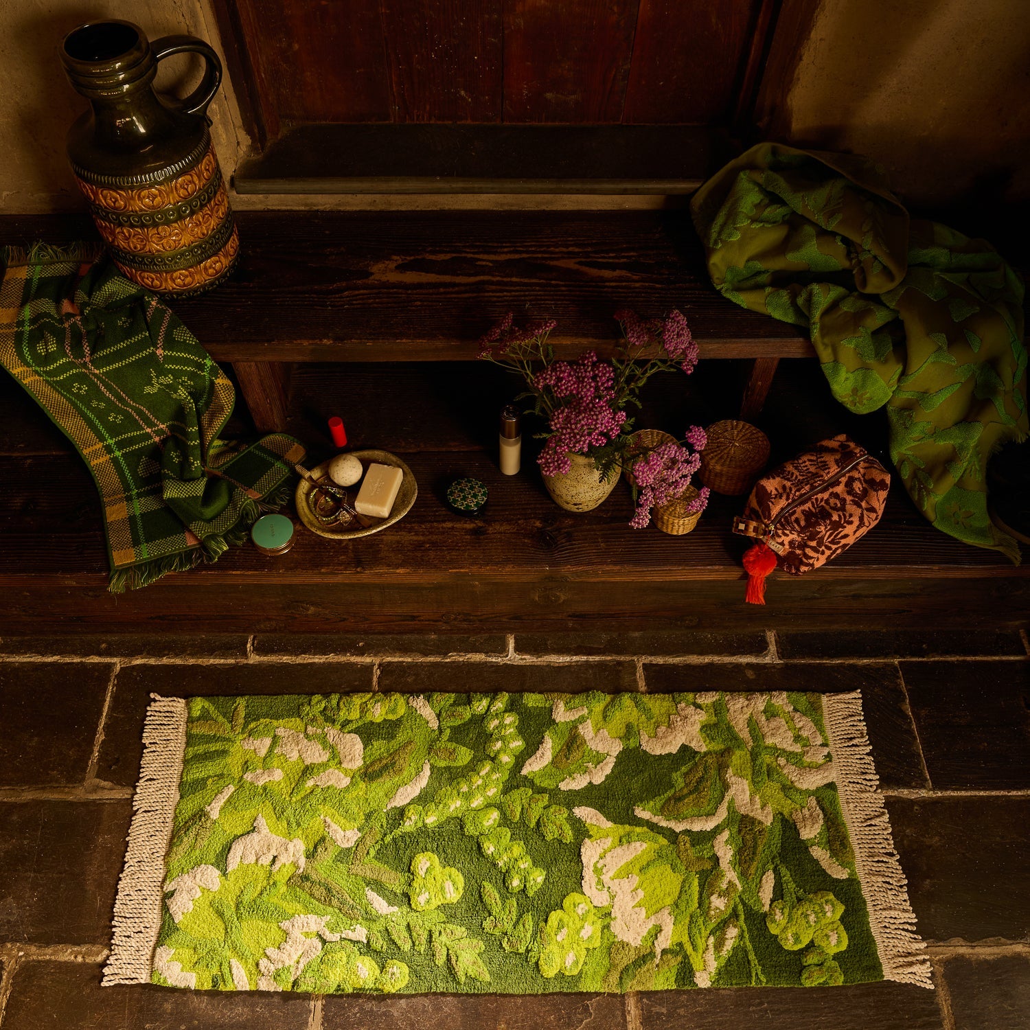 Overhead view of green floral tufted bath runner with cream fringe on stone floor beside a wooden bench