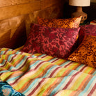 Red floral and orange patterned linen pillowcases stacked on a bed with a multicolour striped sheet