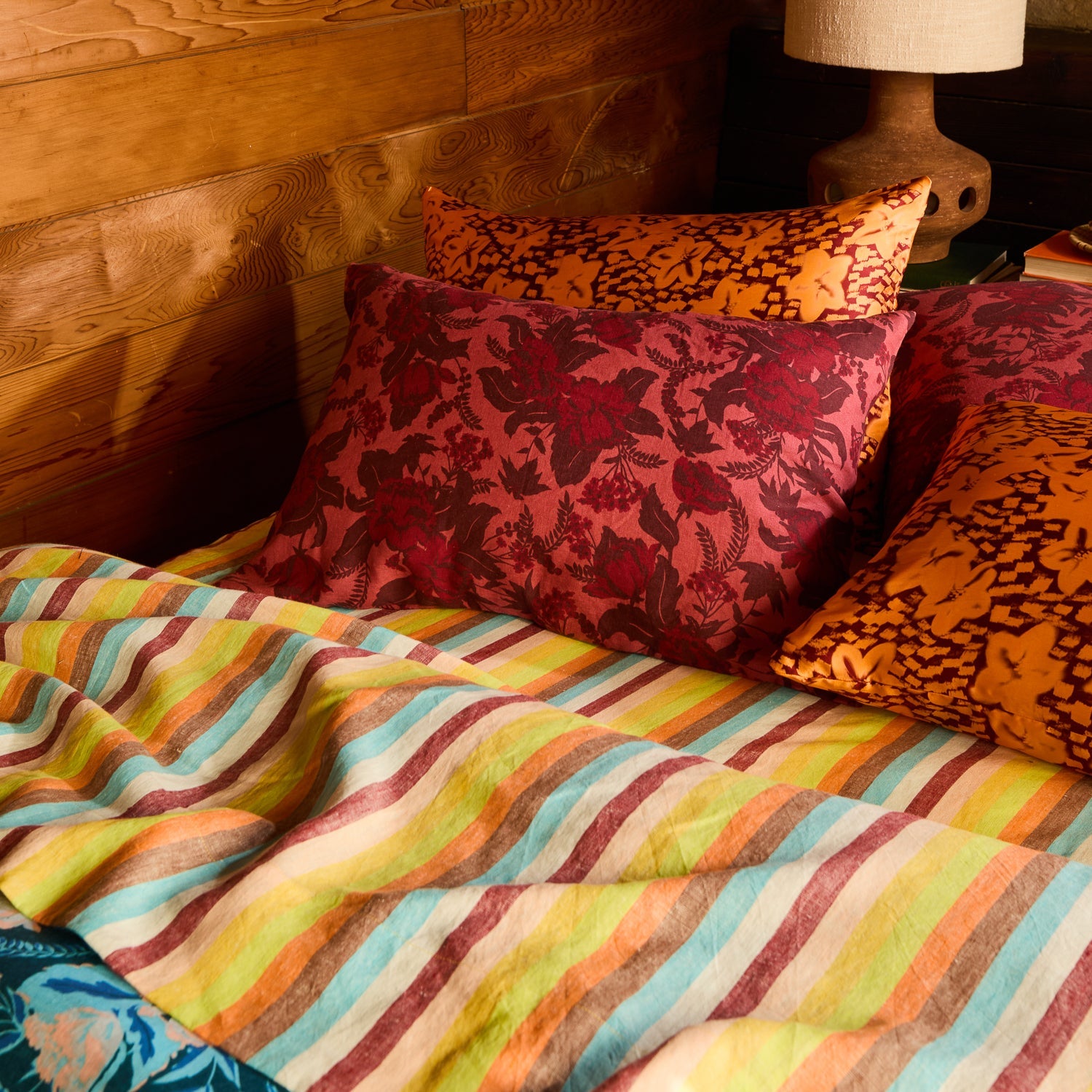 Red floral and orange patterned linen pillowcases stacked on a bed with a multicolour striped sheet