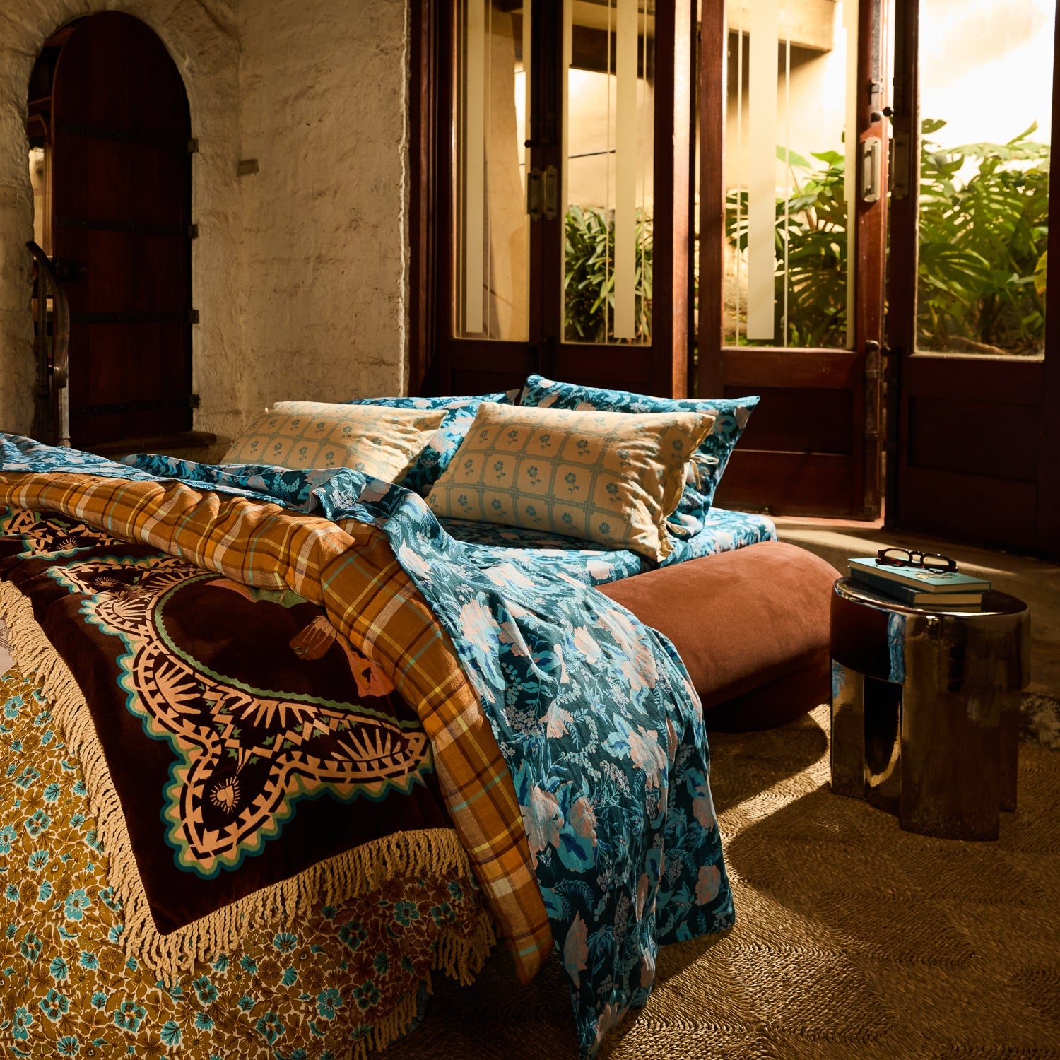 Teal floral linen sheet set styled on a bed in a rustic room with open doors to a tropical garden