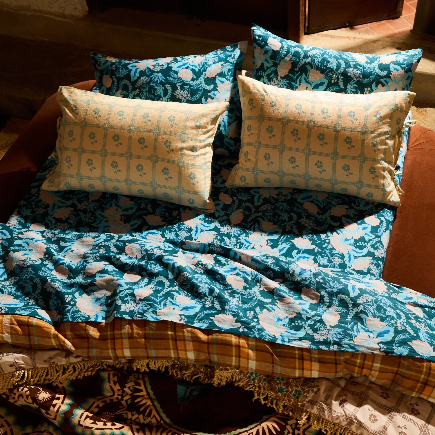 Teal botanical floral linen sheet and pillowcases layered on a sunlit bed with mixed-print bedding