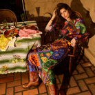 Woman seated in a chair wearing a multicolour patchwork-print maxi dress with a black belt and cowboy boots