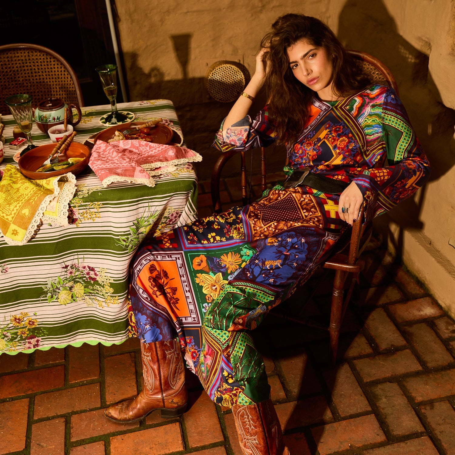 Woman seated in a chair wearing a multicolour patchwork-print maxi dress with a black belt and cowboy boots