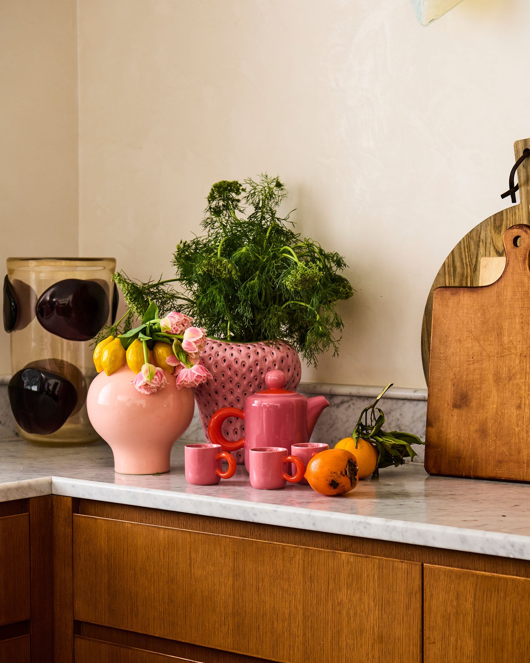Pink ceramic espresso mugs with orange handles arranged on kitchen counter with plants and oranges