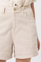 Close-up front detail of cream denim shorts showing heart-shaped gold button, front pocket, and contrast stitching