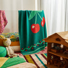Tea Baby Blanket in green knit with red cherry pattern displayed in colorful nursery playroom