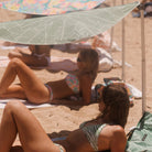 Two women in bikinis lying under a leaf-patterned Swim Shady beach umbrella on a sandy beach.