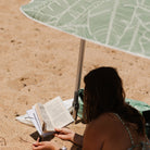 Person reading a book under a green leaf-patterned umbrella on a sandy beach.