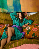 Woman relaxing on colorful floral bedding wearing teal Tropicas Adult Cosy Robe with purple accents