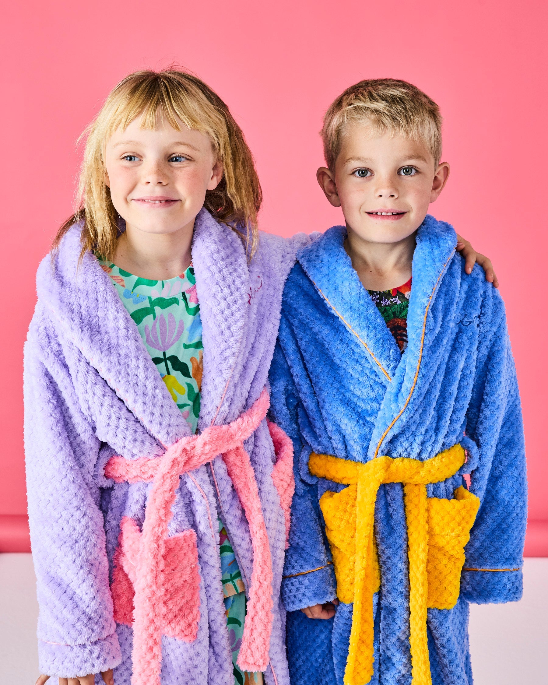 Two children wearing Tulip Kids Cozy Robes in purple and blue with colorful belts on pink background