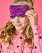 Model wearing Kip & Co Wild Grape Velvet Eye Mask in purple with teal trim on pink background