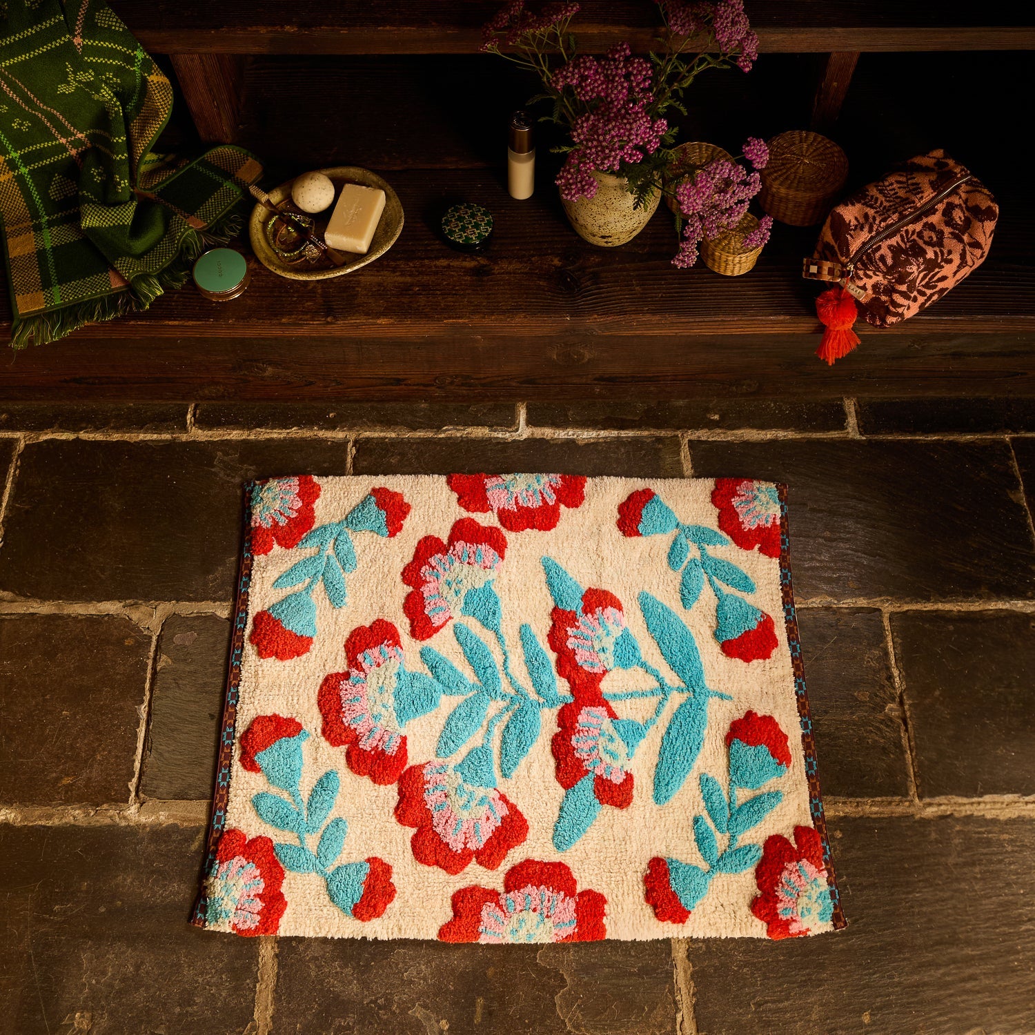 Moonlight Winona Bath Mat with red floral and teal leaf pattern placed on a stone floor below a wooden shelf