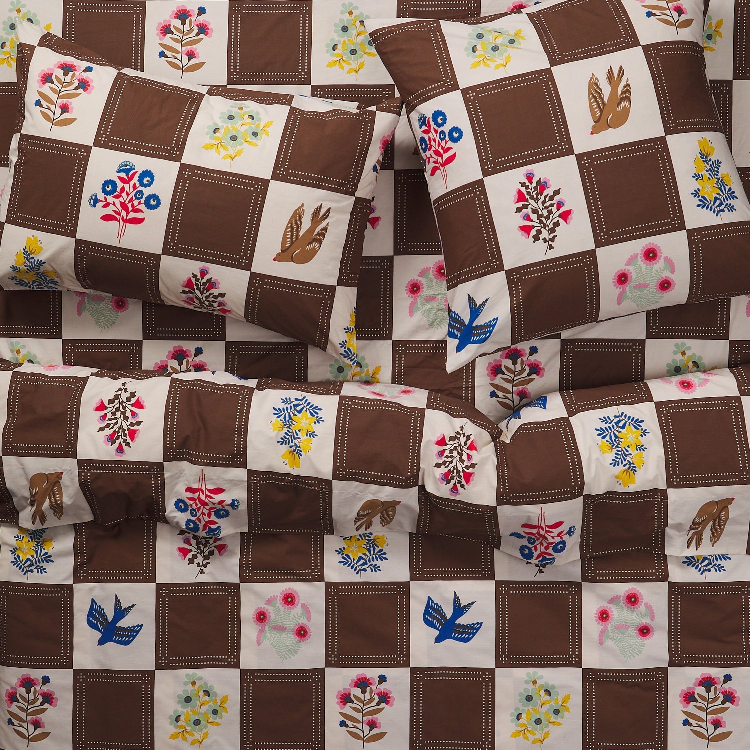 Pair of brown checkerboard cotton pillowcases with colorful folk floral and bird print, stacked on matching bedding
