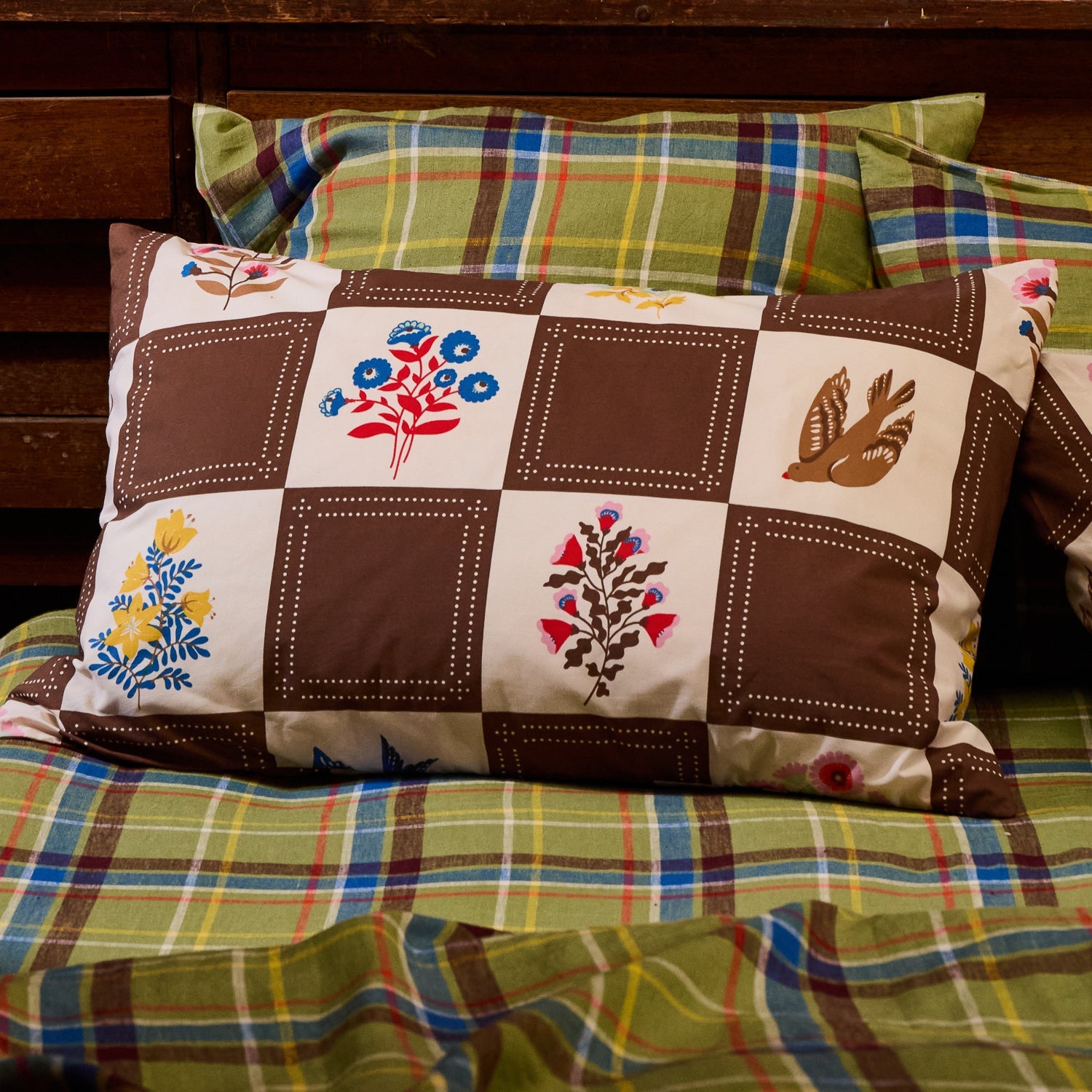 Brown and cream checkerboard cotton pillowcase with folk floral and bird print, styled on a bed with plaid bedding