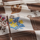 Flat-lay detail of cotton robe fabric showing brown and cream checkered print with bird motif, blue and yellow floral pocket, and dot border