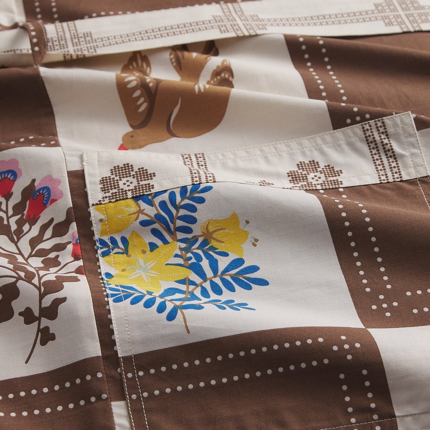 Flat-lay detail of cotton robe fabric showing brown and cream checkered print with bird motif, blue and yellow floral pocket, and dot border