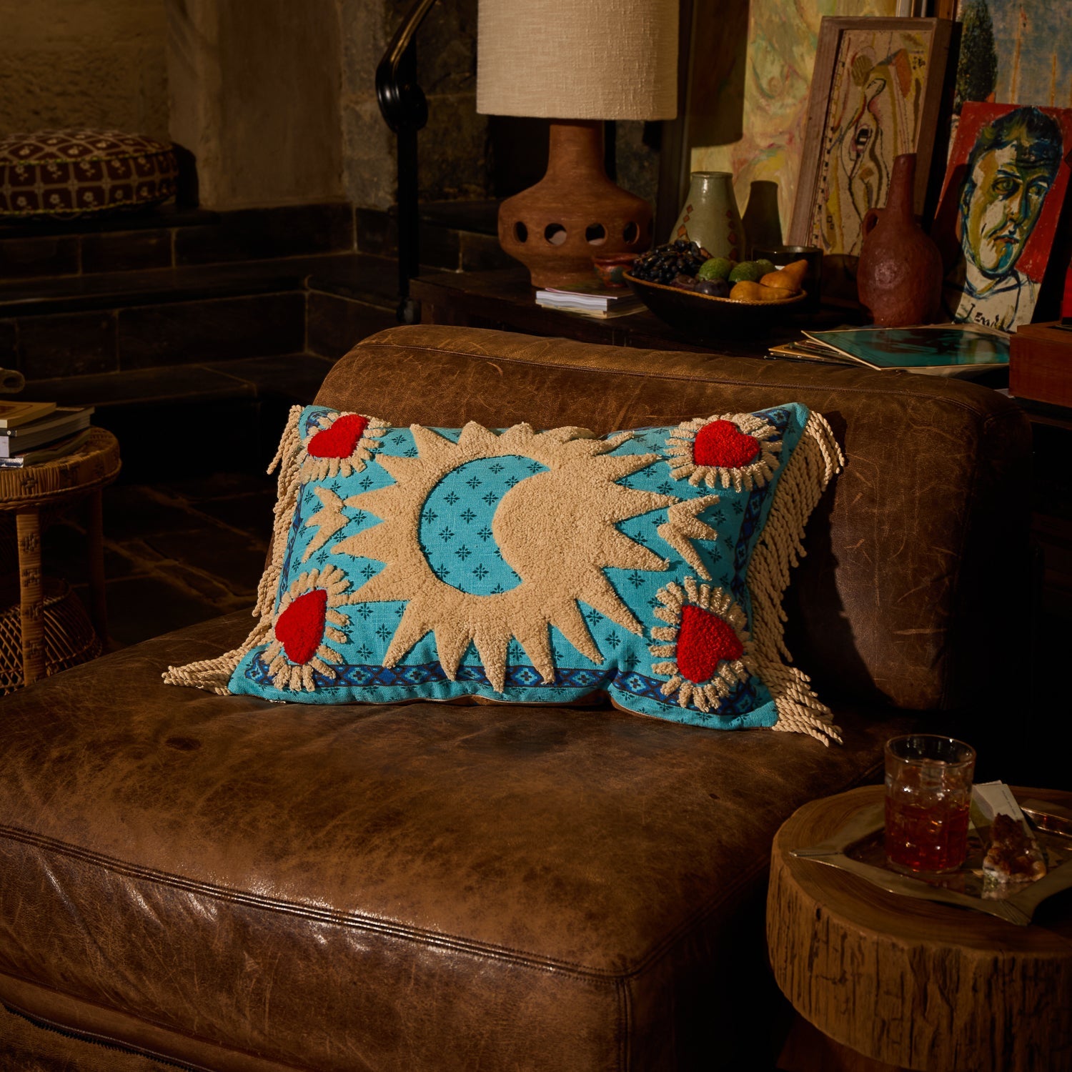 Turquoise reversible tufted cushion with cream sun and moon design and red hearts styled on a brown leather sofa