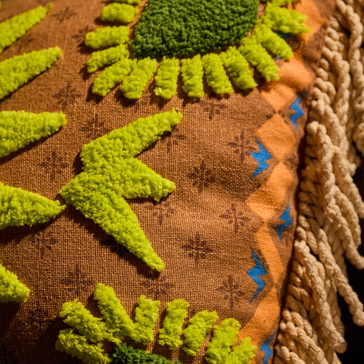 Detail shot of lime green tufted sun motif and rope fringe trim on a brown and orange patterned reversible cushion